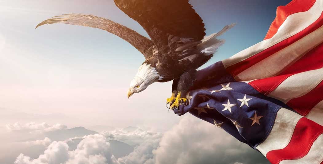 A bald eagle flying over the clouds with an american flag.